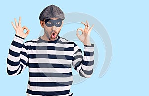 Young handsome man wearing burglar mask looking surprised and shocked doing ok approval symbol with fingers