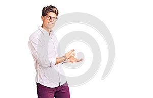 Young handsome man wearing business clothes and glasses inviting to enter smiling natural with open hand