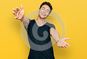 Young handsome man wearing casual black t shirt looking at the camera smiling with open arms for hug