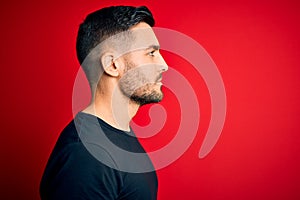 Young handsome man wearing casual black t-shirt standing over isolated red background looking to side, relax profile pose with photo