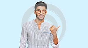 Young handsome man wearing casual clothes and glasses smiling with happy face looking and pointing to the side with thumb up