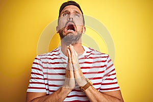 Young handsome man wearing casual red striped t-shirt over yellow isolated background begging and praying with hands together with