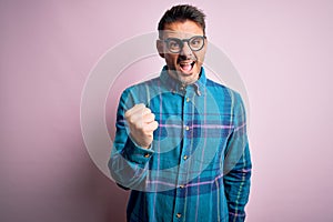 Young handsome man wearing casual shirt and glasses standing over isolated pink background angry and mad raising fist frustrated