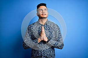 Young handsome man wearing casual shirt standing over isolated blue background begging and praying with hands together with hope