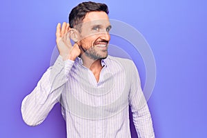 Young handsome man wearing casual striped shirt standing over isolated purple background smiling with hand over ear listening and