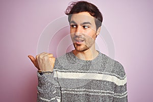 Young handsome man wearing casual striped sweater standing over isolated pink background pointing and showing with thumb up to the