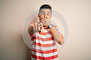 Young handsome man wearing casual striped t-shirt standing over isolated white background laughing at you, pointing finger to the