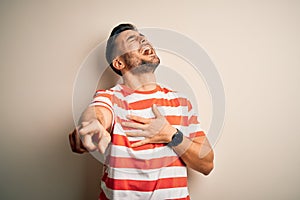 Young handsome man wearing casual striped t-shirt standing over isolated white background laughing at you, pointing finger to the