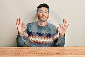 Young handsome man wearing casual sweater sitting on the table relax and smiling with eyes closed doing meditation gesture with