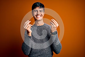 Young handsome man wearing casual sweater standing over isolated orange background doing money gesture with hands, asking for