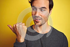 Young handsome man wearing casual sweater standing over isolated yellow background pointing and showing with thumb up to the side