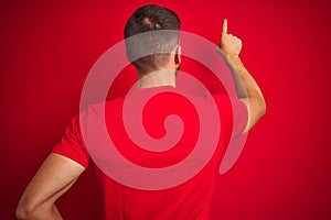 Young handsome man wearing casual t-shirt over red isolated background Posing backwards pointing ahead with finger hand