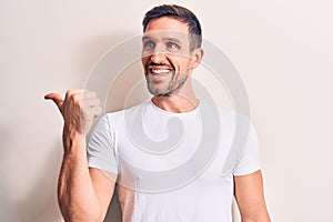 Young handsome man wearing casual t-shirt standing over isolated white background pointing thumb up to the side smiling happy with