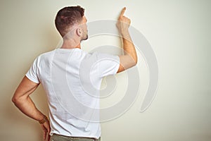Young handsome man wearing casual white t-shirt over isolated background Posing backwards pointing ahead with finger hand