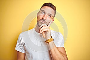 Young handsome man wearing casual white t-shirt over yellow isolated background with hand on chin thinking about question, pensive