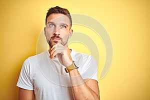 Young handsome man wearing casual white t-shirt over yellow isolated background with hand on chin thinking about question, pensive