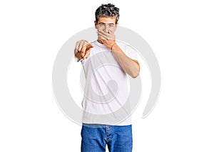 Young handsome man wearing casual white tshirt laughing at you, pointing finger to the camera with hand over mouth, shame