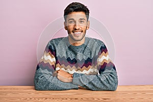 Young handsome man wearing casual winter sweater sitting on the table with a happy and cool smile on face