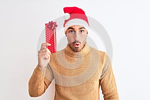 Young handsome man wearing christmas hat and holding gift over isolated background scared in shock with a surprise face, afraid