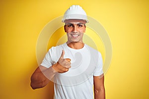 Young handsome man wearing construction helmet over yellow isolated background doing happy thumbs up gesture with hand