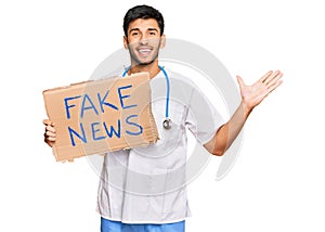 Young handsome man wearing doctor uniform holding fake news banner celebrating victory with happy smile and winner expression with