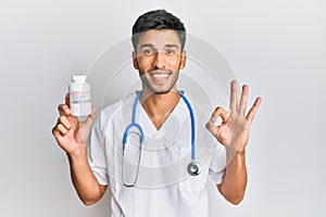 Young handsome man wearing doctor uniform holding presciption pills doing ok sign with fingers, smiling friendly gesturing