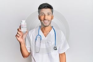 Young handsome man wearing doctor uniform holding presciption pills looking positive and happy standing and smiling with a