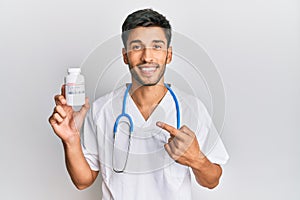 Young handsome man wearing doctor uniform holding presciption pills smiling happy pointing with hand and finger
