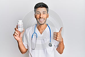 Young handsome man wearing doctor uniform holding presciption pills smiling happy and positive, thumb up doing excellent and