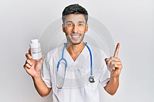 Young handsome man wearing doctor uniform holding presciption pills smiling with an idea or question pointing finger with happy