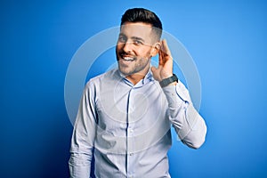 Young handsome man wearing elegant shirt standing over isolated blue background smiling with hand over ear listening an hearing to
