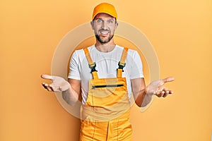 Young handsome man wearing handyman uniform over yellow background smiling cheerful with open arms as friendly welcome, positive