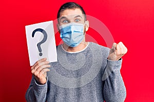 Young handsome man wearing medical mask holding question mark screaming proud, celebrating victory and success very excited with