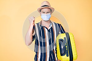 Young handsome man wearing medical mask holding suitcase annoyed and frustrated shouting with anger, yelling crazy with anger and