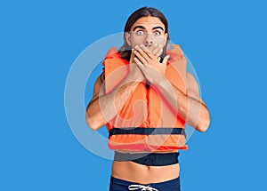 Young handsome man wearing nautical lifejacket shocked covering mouth with hands for mistake