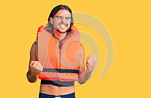 Young handsome man wearing nautical lifejacket very happy and excited doing winner gesture with arms raised, smiling and screaming