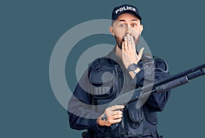 Young handsome man wearing police uniform holding shotgun covering mouth with hand, shocked and afraid for mistake
