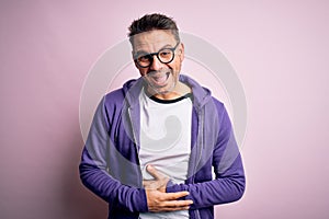 Young handsome man wearing purple sweatshirt and glasses standing over pink background smiling and laughing hard out loud because