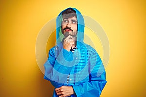 Young handsome man wearing rain coat with hood standing over isolated yellow background with hand on chin thinking about question,