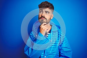 Young handsome man wearing rain coat standing over isolated blue background with hand on chin thinking about question, pensive