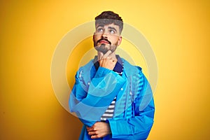 Young handsome man wearing rain coat standing over isolated yellow background with hand on chin thinking about question, pensive