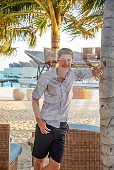 Young handsome man wearing shirt posing near palm tree at the beach at the tropical island luxury resort