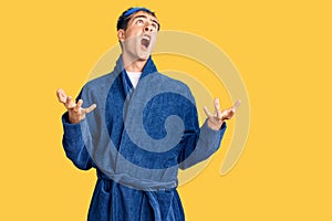 Young handsome man wearing sleep mask and bathrobe crazy and mad shouting and yelling with aggressive expression and arms raised