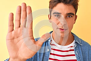 Young handsome man wearing striped t-shirt and denim shirt over isolated yellow background with open hand doing stop sign with