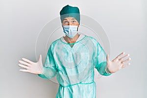 Young handsome man wearing surgeon uniform and medical mask crazy and mad shouting and yelling with aggressive expression and arms