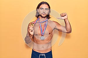 Young handsome man wearing swimwear and medals with angry face, negative sign showing dislike with thumbs down, rejection concept