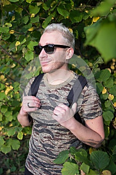 A young handsome man wearing t-shirt and black sunglasses on background of green and yellow leaves.