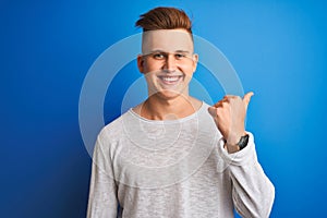 Young handsome man wearing white casual t-shirt standing over isolated blue background pointing and showing with thumb up to the