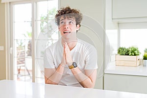 Young handsome man wearing white t-shirt at home begging and praying with hands together with hope expression on face very