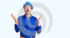 Young handsome man wearing worker uniform and hardhat shouting angry out loud with hands over mouth